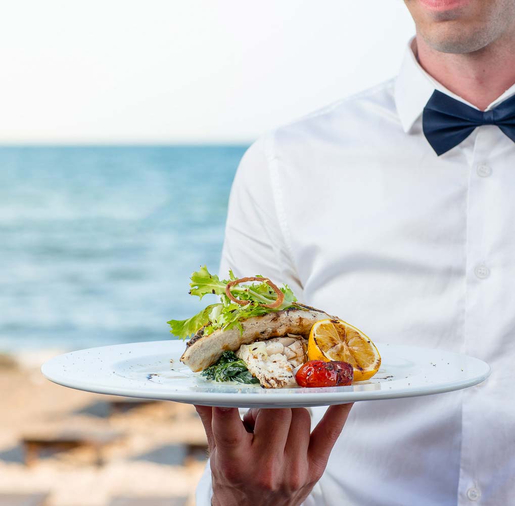 waiter holding grilled fish with lemon tomato creamy herbs seaside restaurant